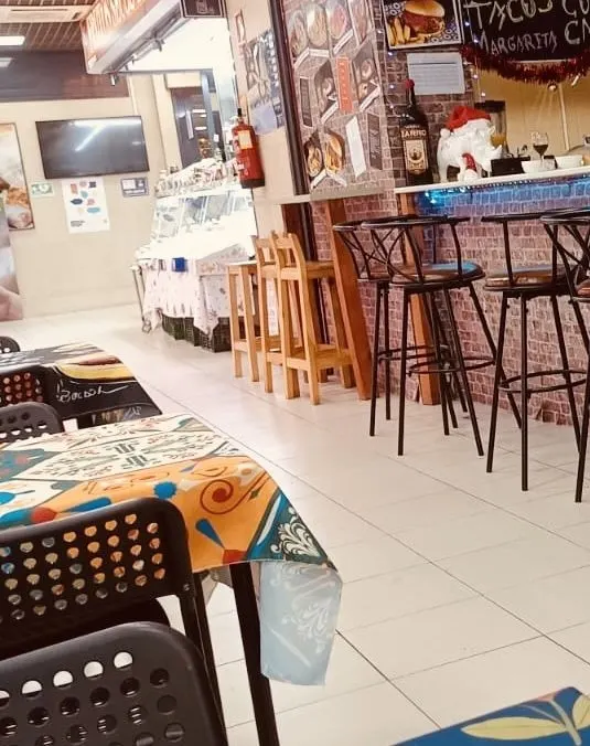 Barra del puesto con taburetes altos, pizarra de tacos al fondo y mesas con mantel de colores